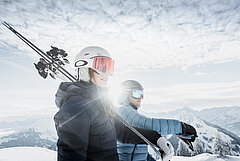 Two skiers carry their skis in the sunshine, with a beautiful landscape behind them.