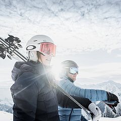Two skiers carry their skis in the sunshine, with a beautiful landscape behind them.