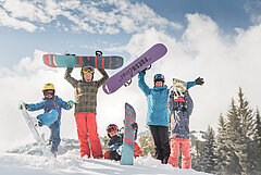 Eine Gruppe von farbig gekleideten Kindern mit ihren Snowboards und der blau gekleideten Snowboardlehrerin vor verschneiter Winterlandschaft.