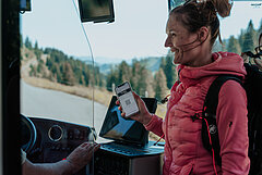Eine Frau mit rosa Daunenjacke und schwarzem Rucksack zeigt dem Buschauffeur auf dem Smartphone ihr Busticket.