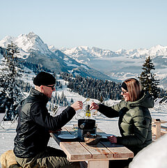 Zwei Personen essen Fondue im Freien vor verschneiten Berggipfeln.