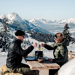 Zwei Personen essen Fondue im Freien vor verschneiten Berggipfeln.