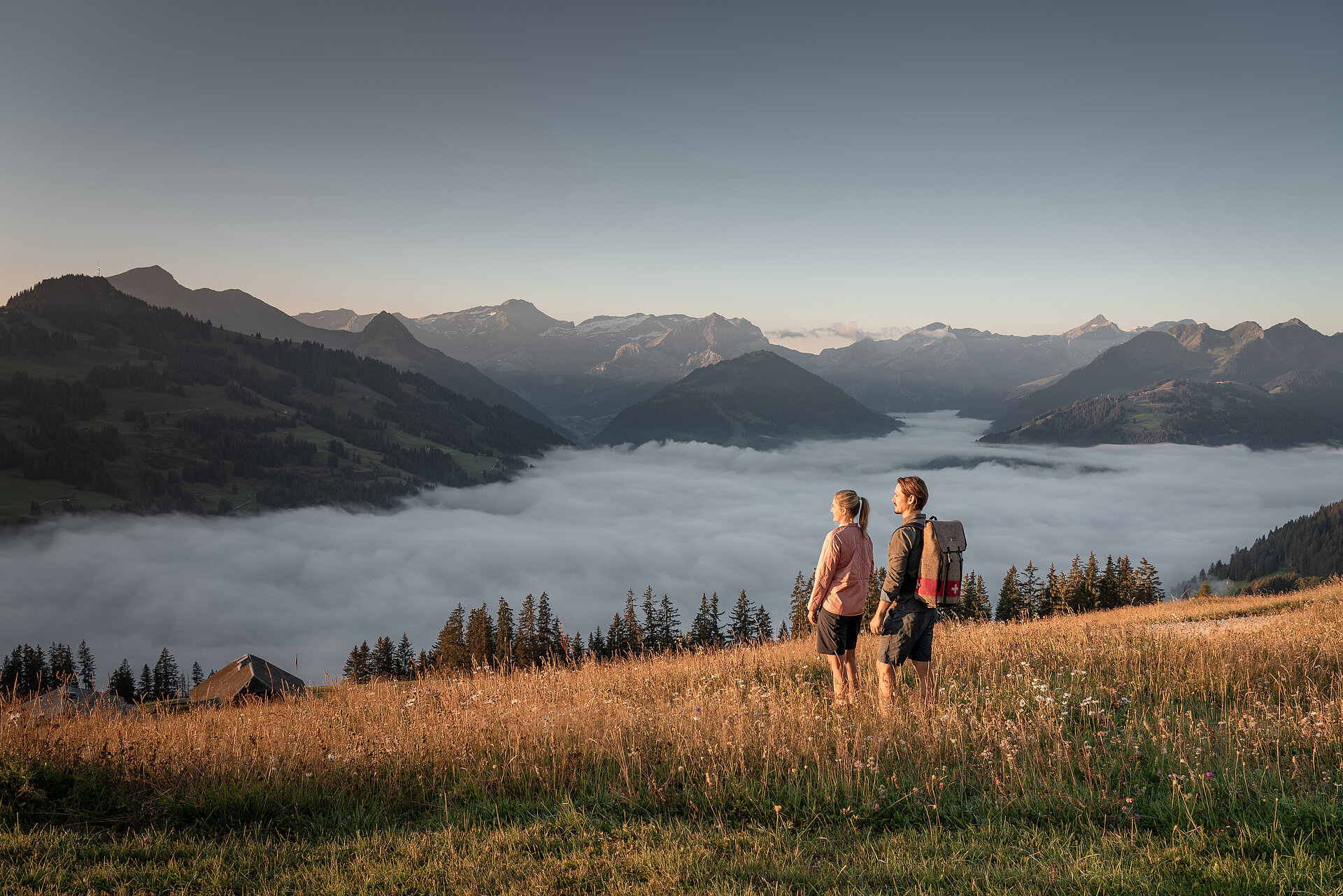 Zwei Personen, welche in einer Wiese auf einem Berg stehen und den Sonnenuntergang in einem schönen Bergpanorama betrachten.