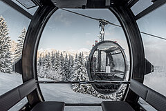 The photo is from a modern 10-seater gondola lift. The gondola has black leather benches and lots of glass. The design is oval and looks futuristic. Outside the gondola, another gondola is crossing with two people inside. Outside, the landscape is covered in snow and only on the mountain peaks can you see the sun.