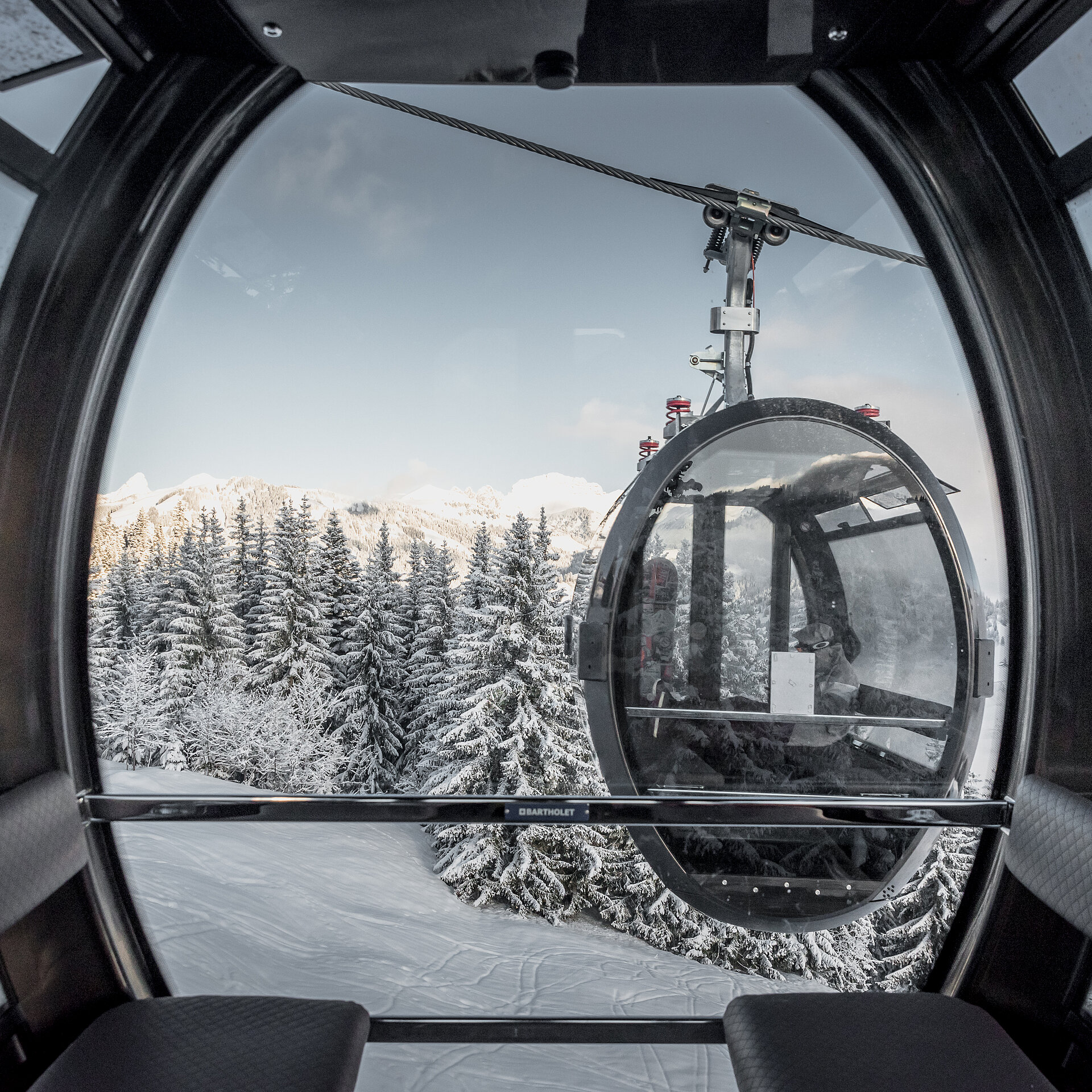 Das Foto stammt aus einer modernen 10er-Gondelbahn. Die Gondelbahn hat schwarze Lederbänke und viel Glas. Das Design ist oval und wirkt futuristisch. Ausserhalb der Gondel kreuzt eine andere Gondel mit zwei Personen darin. Draussen ist die Landschaft verschneit und nur auf den Bergspitzen sieht man die Sonne.