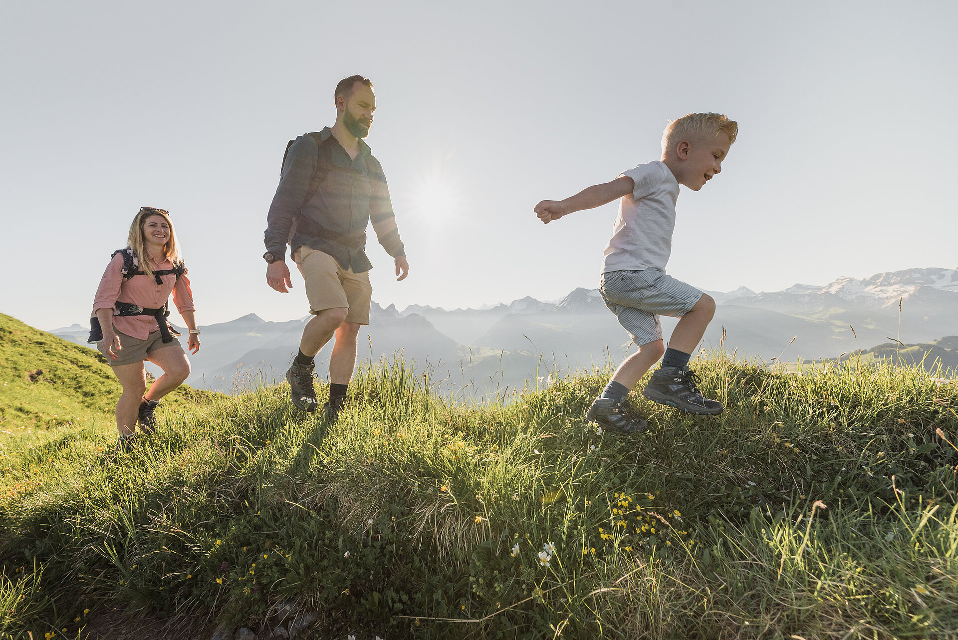 Familie bestehend aus drei Personen beim Wandern über grüne Wiesen bei sommerlichem Wetter.