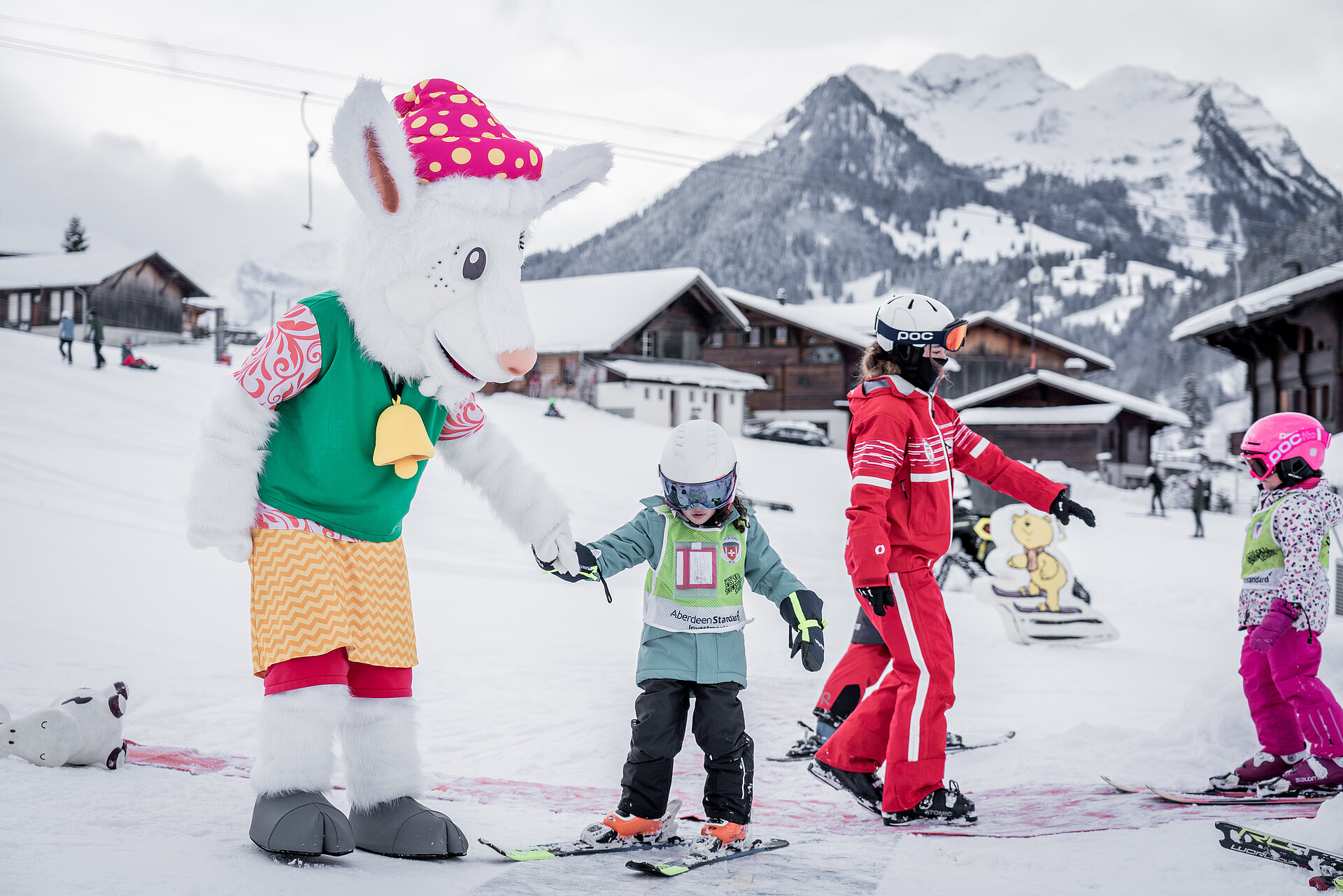 Das Maskottchen Saani im Kinderland der Skischule Gstaad mit einer rot gekleideten Skilehrerin und zwei kleinen Kindern auf Ski.