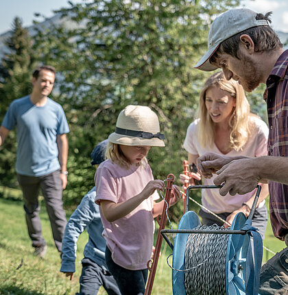 Zwei Kinder helfen einem Bauern mit dem Zaun aufstellen. Die Eltern stehen mit Gabeln etwas weiter hinten im Bild. Es ist sonnig und sie stehen auf einer grünen Wiese.