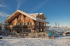 A picturesque mountain cabin in the snow with a clear blue sky. In the foreground, skiers and a gondola can be seen.