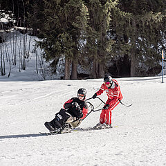 Ein Skilehrer in roter Skilehrerbekleidung führte eine Person in einem Sitzski auf der Piste