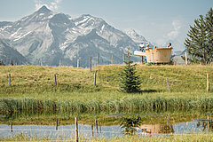Im Vordergrund ein kleines Bergseeli, dahinter ein XXL Fondue-Caquelon und im Hintergrund ein Bergpanorama.
